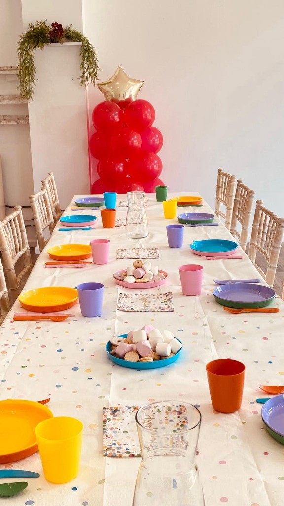 Long celebration table with wooden chairs and colourful tableware beside windows overlooking the countryside.
